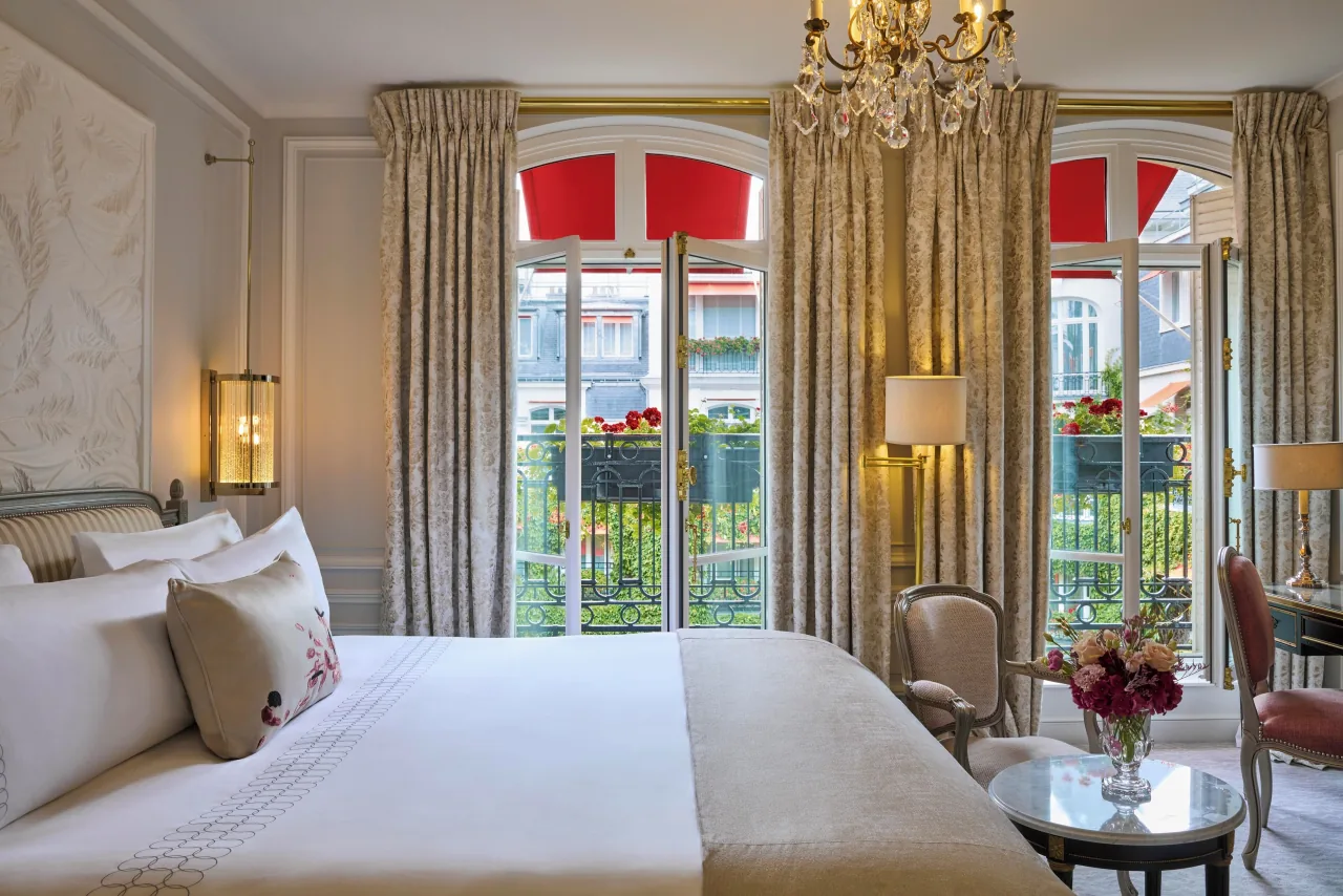 A bedroom with a wide white bed, tall curtains and French windows under red awnings, opening onto a balcony with red geraniums