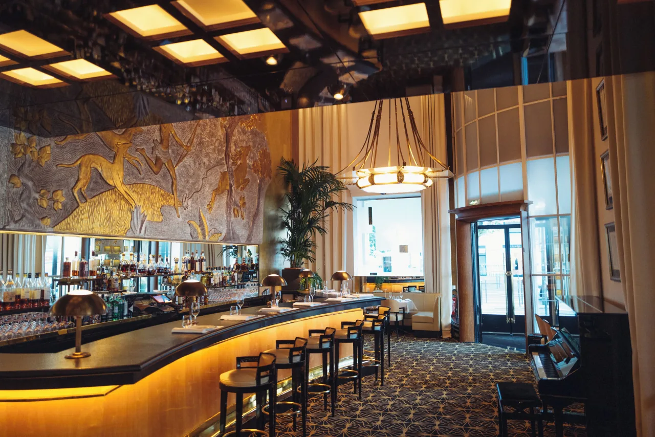 An Art Deco room with a curved, lit bar and high stools below a gilded hunting fresco, a coffered ceiling and a palm
