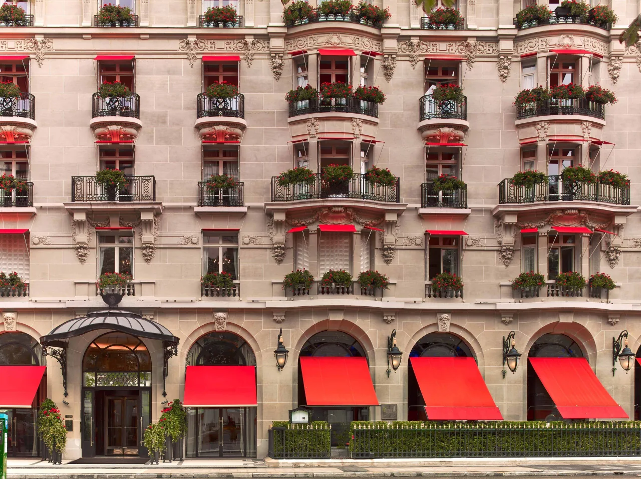 The stone façade on avenue Montaigne: wrought-iron balconies with red geraniums, a red awning at each window, arched ground-floor bays with red awnings and a glass canopy over the entrance