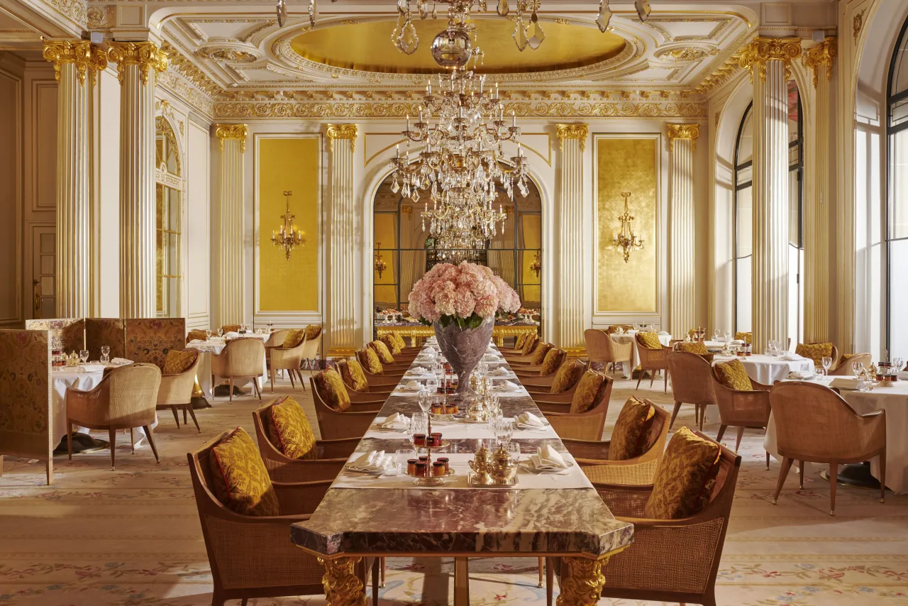 A gilded dining room with columns and a crystal chandelier, a long marble table set with pink roses and wicker armchairs