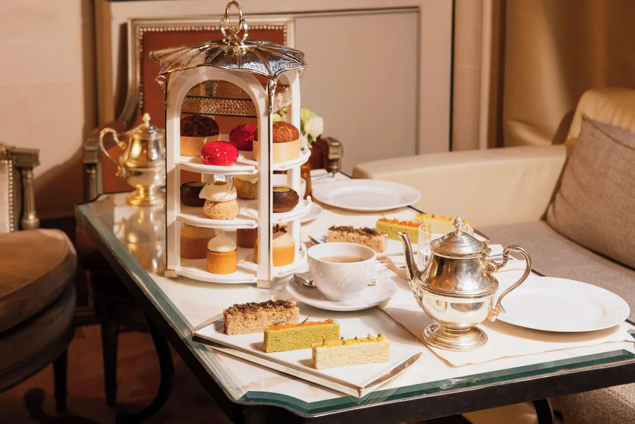 Afternoon tea on a glass table: a silver tiered stand of pastries, a silver teapot, a cup of tea and slices of cake