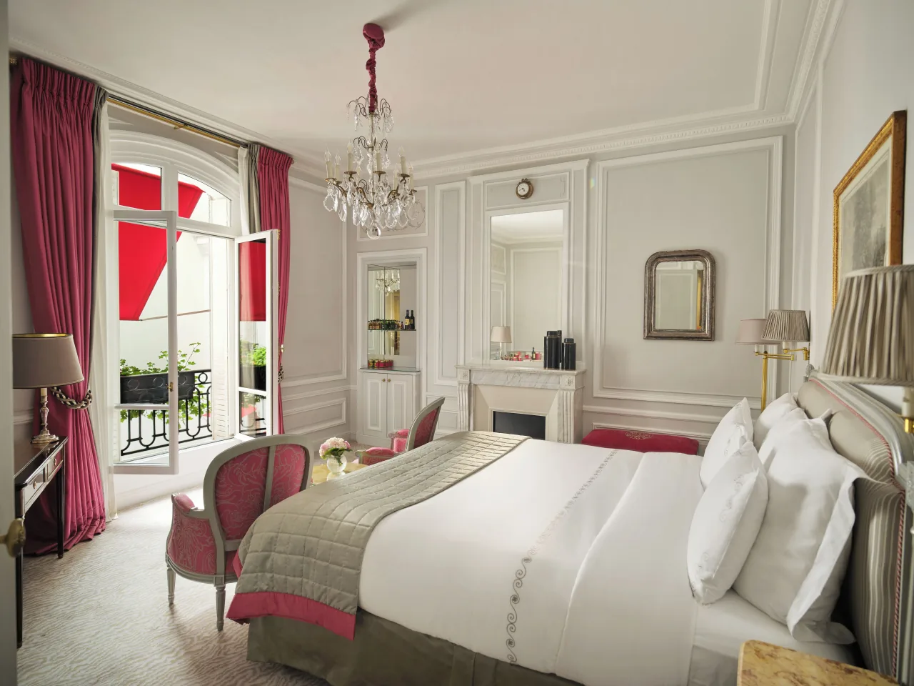 A pale panelled bedroom with a white bed, pink armchairs, a marble fireplace, a crystal chandelier and a French window onto a small balcony