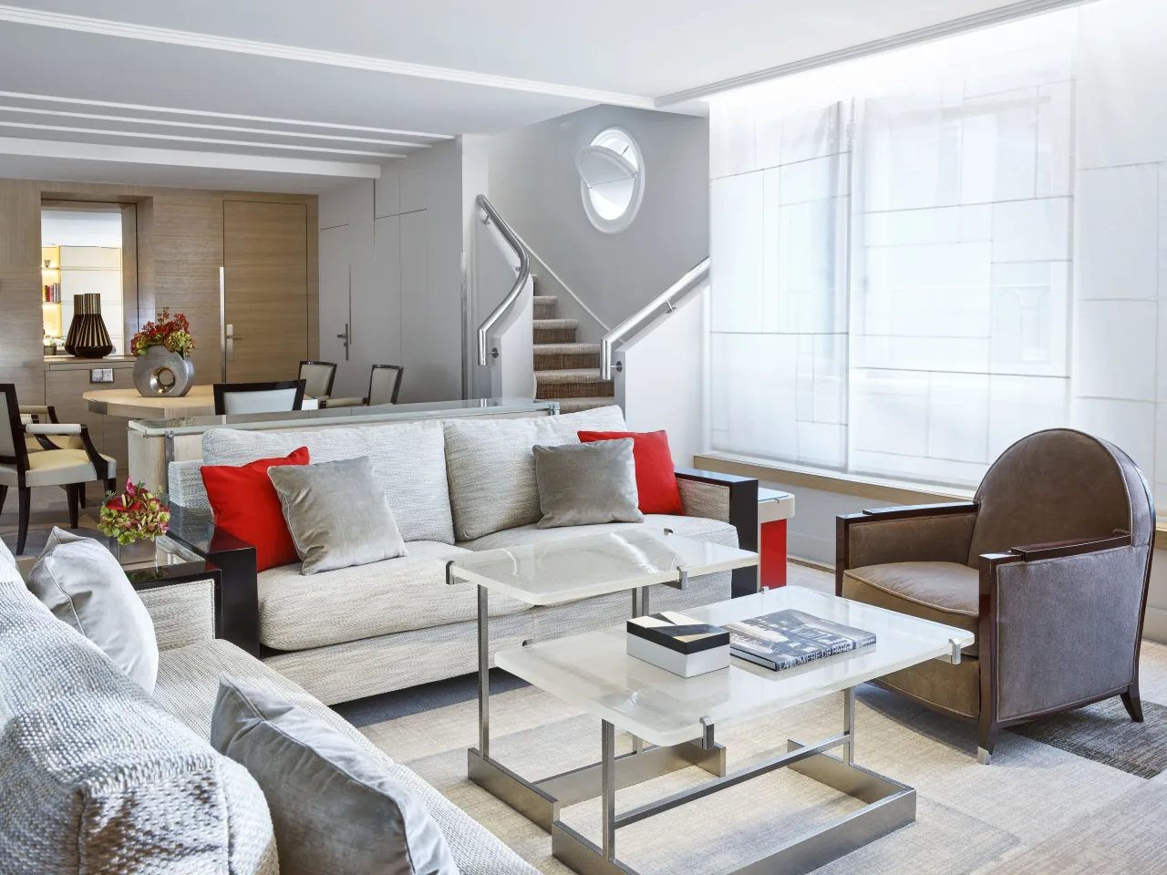 A bright Art Deco living room with pale sofas and red cushions, a glass coffee table, a leather armchair and a staircase leading up
