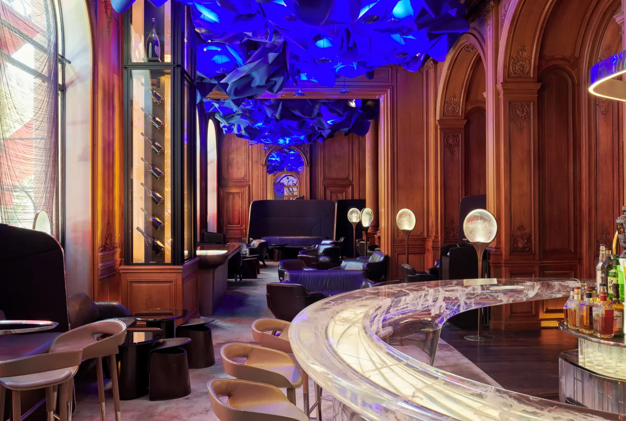A bar with a curved translucent counter under a deep blue ceiling installation, in a wood-panelled room