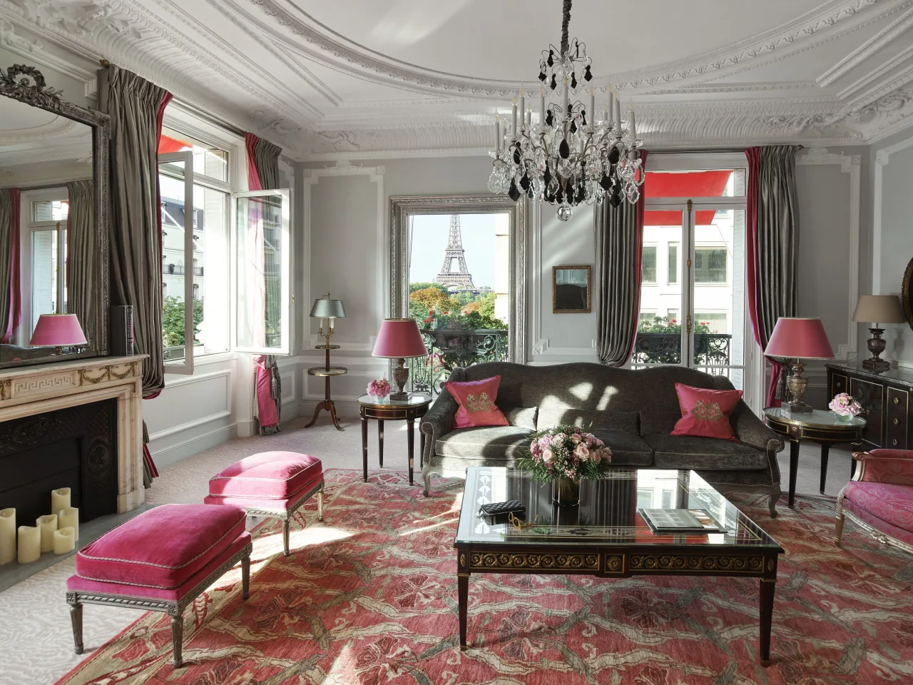 A suite living room with a grey sofa, pink stools, a fireplace, a crystal chandelier and a patterned rug; the Eiffel Tower appears through the central window