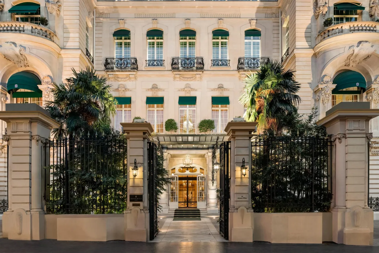The stone façade of Shangri-La Paris behind its gates and palm trees, with the lit entrance