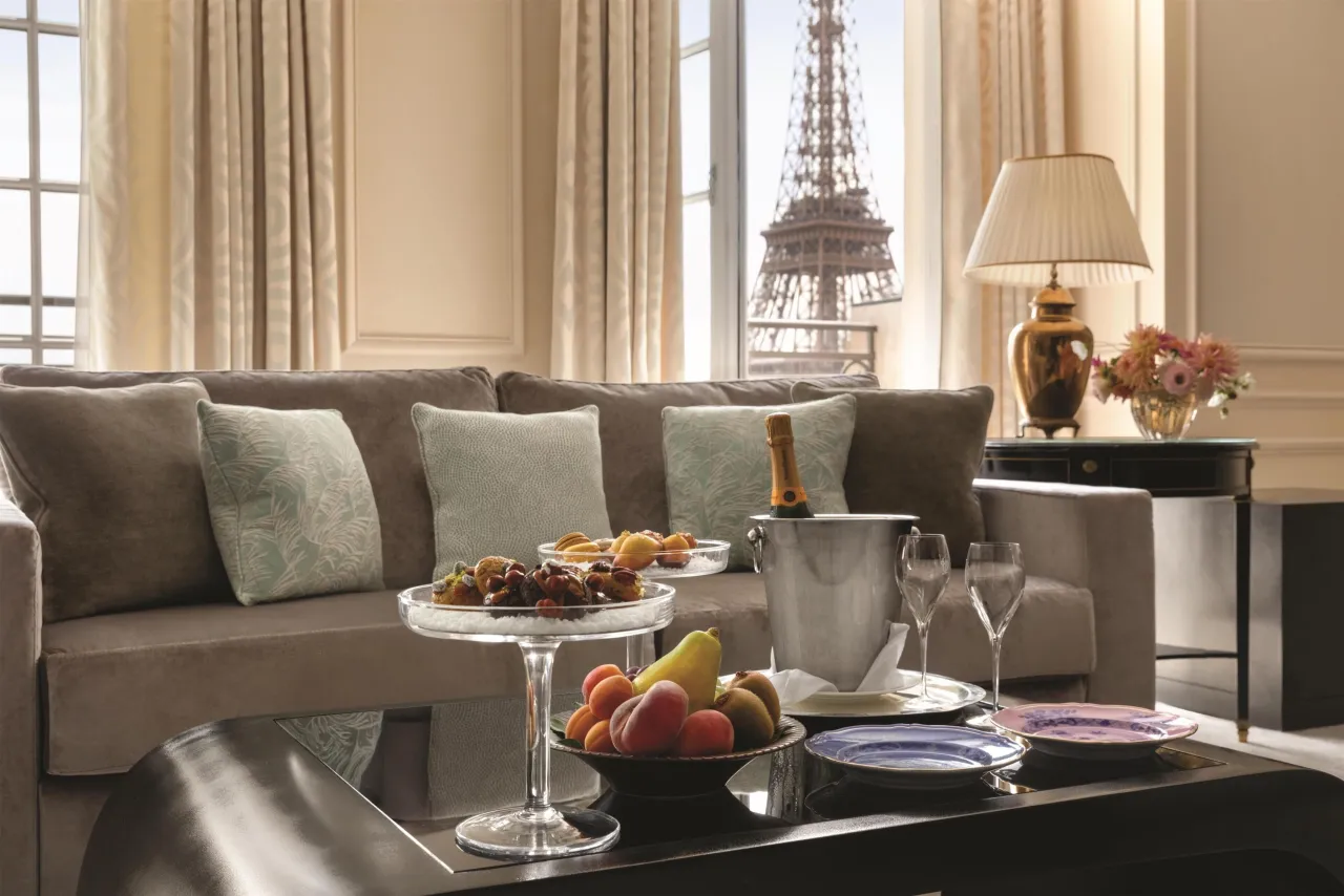 A suite living room with sofas and a table set with fruit and champagne, the Eiffel Tower in the window