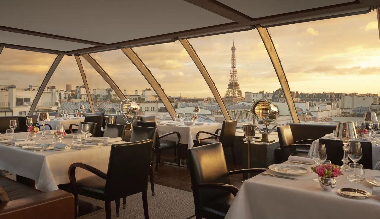 Un restaurant sous une verrière inclinée au coucher du soleil, tables dressées face aux toits de Paris et à la tour Eiffel