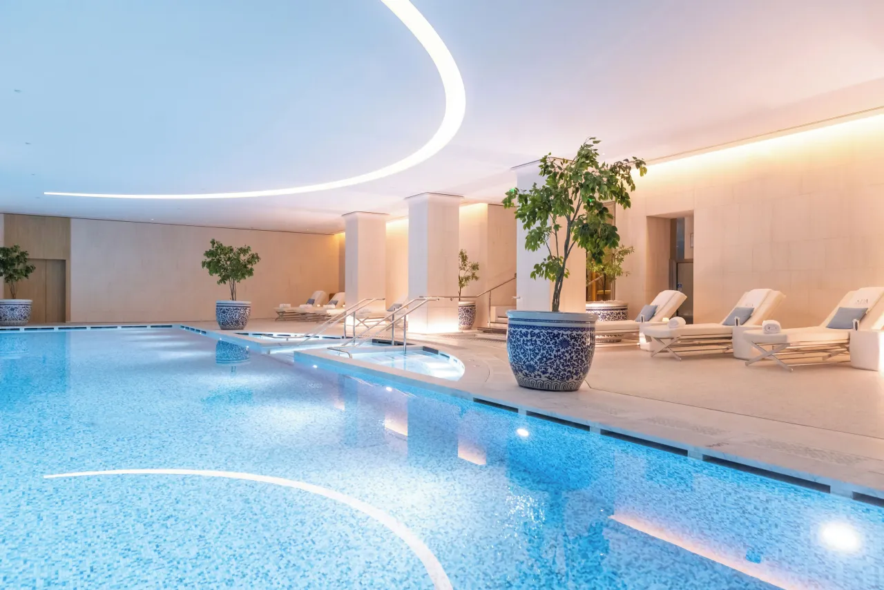 Une longue piscine intérieure en mosaïque bleue sous une ligne lumineuse courbe, chaises longues blanches sur un côté