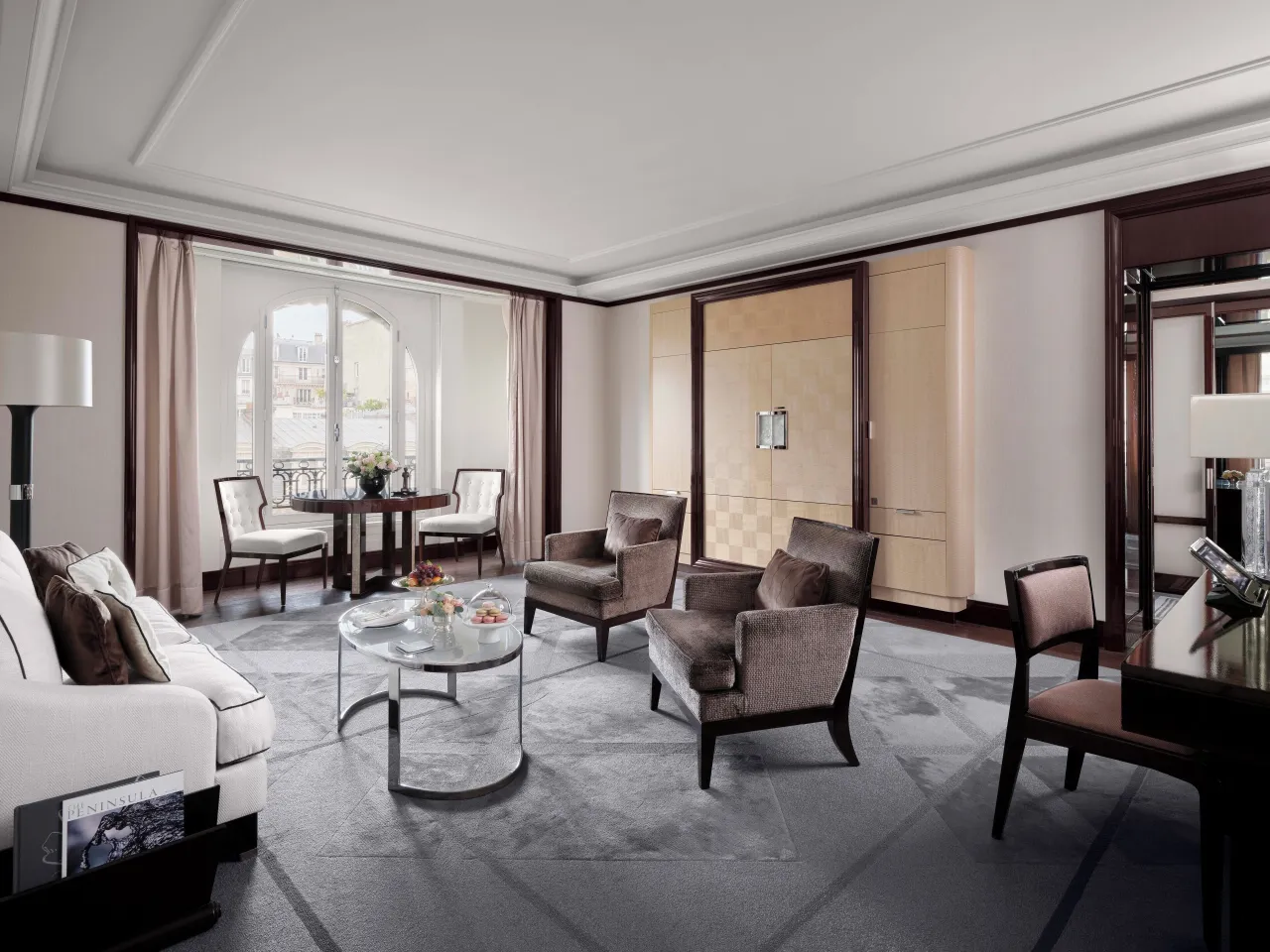 Le salon d’une suite en gris et crème, canapé, deux fauteuils, table en verre et table à manger près de la fenêtre