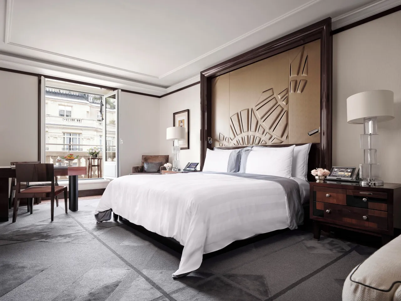 Une chambre au lit blanc adossé à une tête de lit sculptée en éventail, un bureau et une porte-fenêtre ouverte