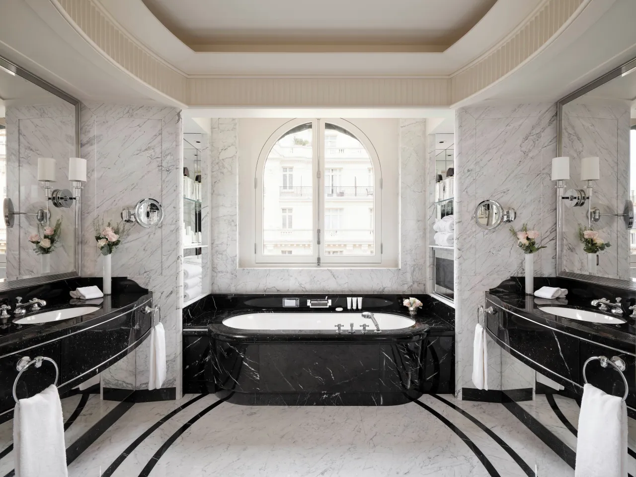 Une salle de bains de marbre blanc, baignoire de marbre noir sous une fenêtre cintrée et deux vasques noires