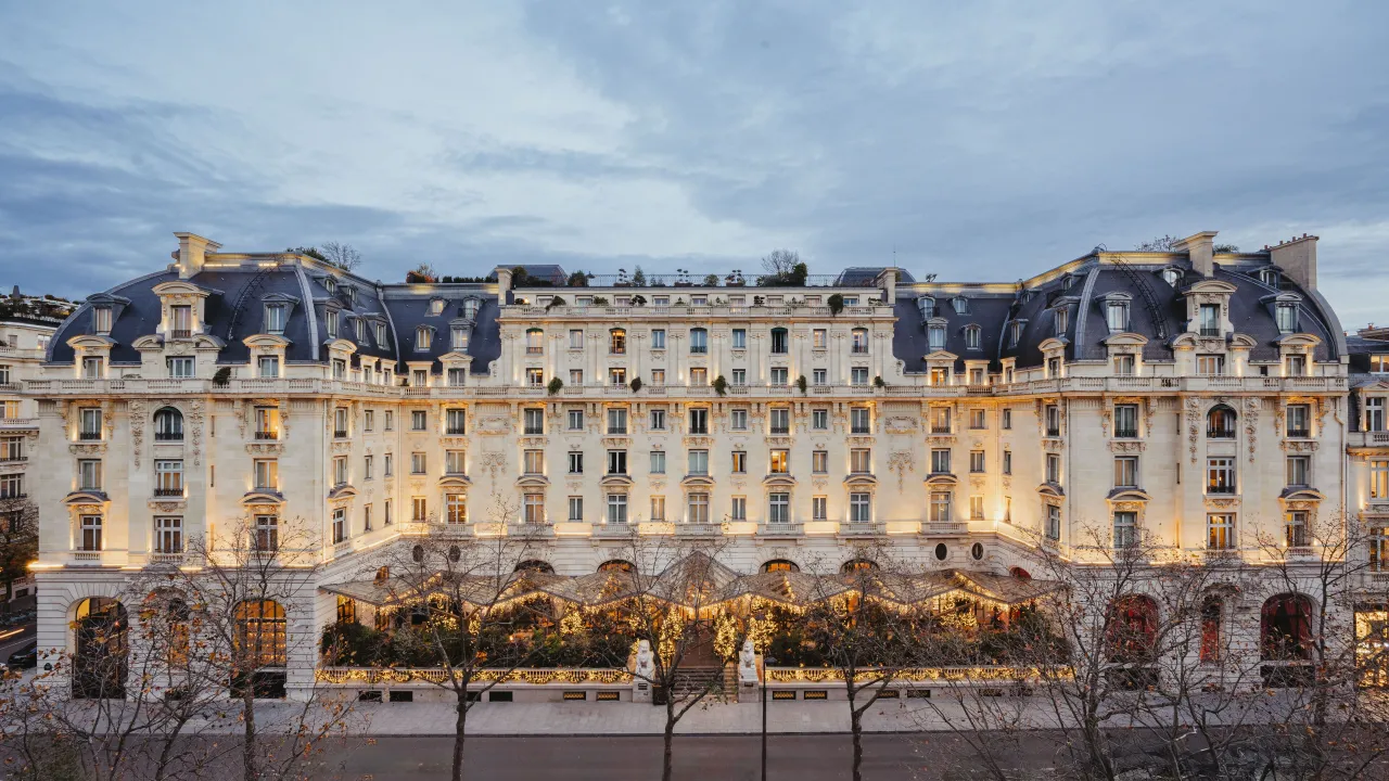 La longue façade de pierre du Peninsula Paris au crépuscule, toits d’ardoise, dômes et rez-de-chaussée éclairé le long de l’avenue