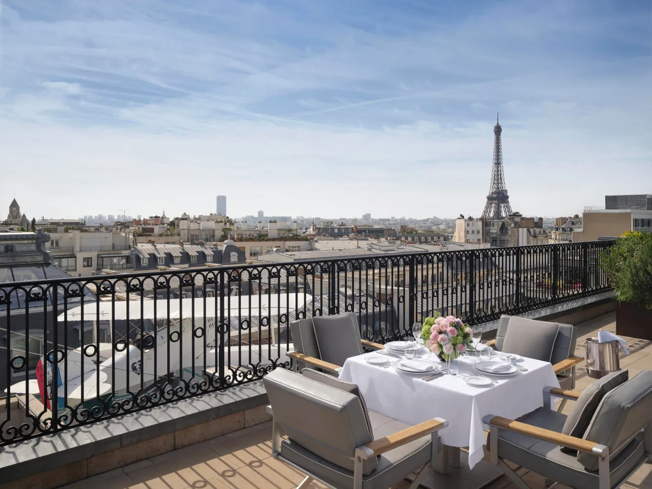 Une table pour quatre dressée sur une terrasse à garde-corps de fer forgé, la tour Eiffel au-dessus des toits