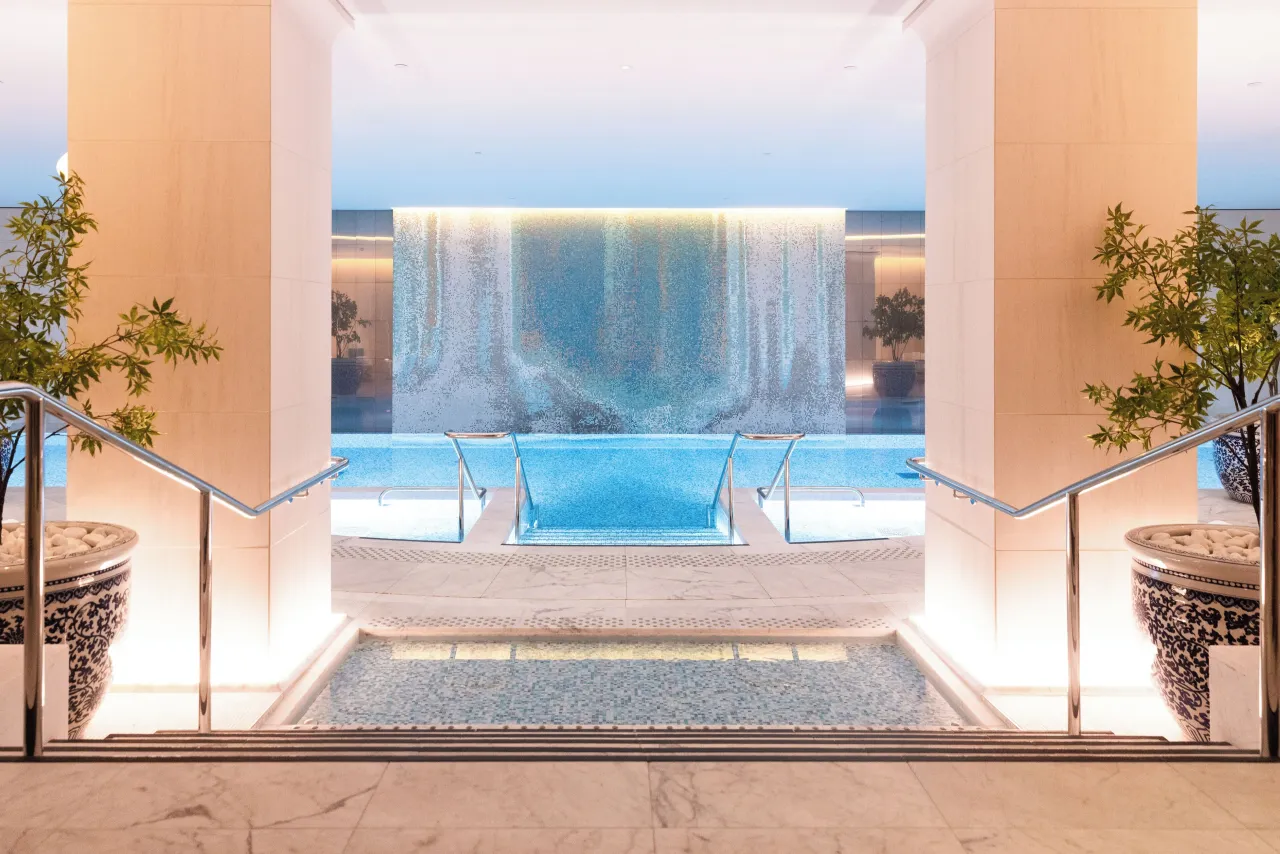 Des marches vers une piscine intérieure entre deux colonnes claires, un mur d’eau éclairé au fond et des plantes en pots bleu et blanc
