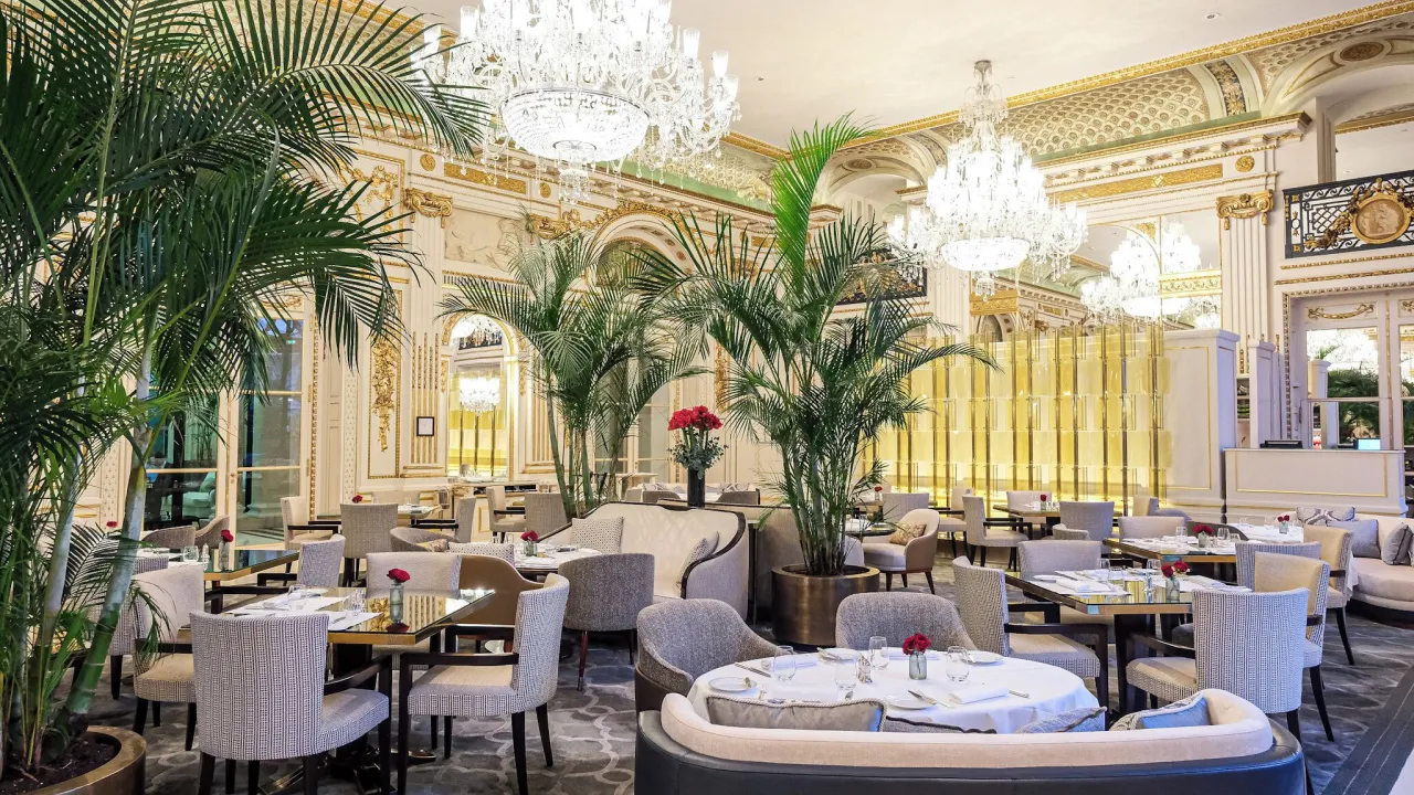 Un restaurant aux moulures dorées, lustres de cristal, palmiers et fauteuils gris autour des tables dressées