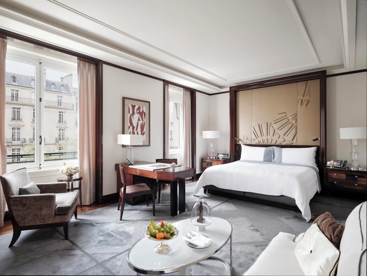 Une chambre avec coin salon, table basse en verre et coupe de fruits, bureau et hautes fenêtres